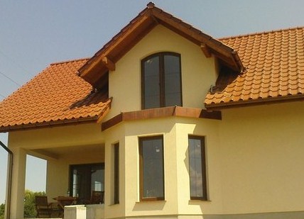 Jasnożółty dom jednorodzinny z brązową dachówką, drewnianymi elementami wykończeniowymi i trzema pionowymi oknami w wykuszu.